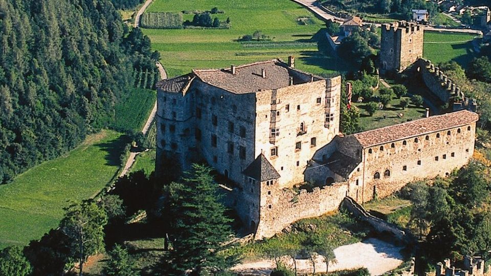Veduta aerea di un castello medievale in pietra con torri e mura merlate, situato su un rilievo tra campi coltivati e un’area boschiva. Il complesso comprende un grande edificio principale e un lungo corpo fortificato collegato a una torre isolata.