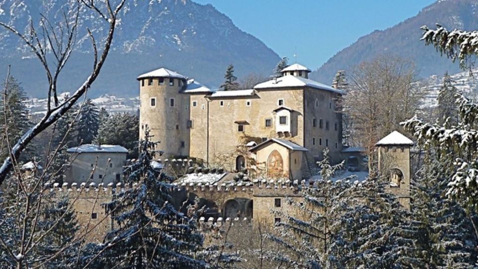 Castello in pietra con mura merlate e torri cilindriche, con tetti coperti di neve, immerso in un bosco innevato. Sullo sfondo si vedono montagne e pendii alpini sotto un cielo sereno.