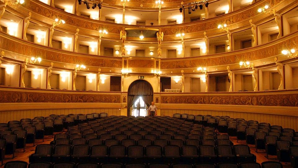 Interno di un teatro storico con platea di sedie scure e tre ordini di palchi curvilinei decorati in tonalità oro, illuminati da lampadari e appliques. Al centro si vede un grande arco con tendaggi che conduce a un ingresso sul fondo.