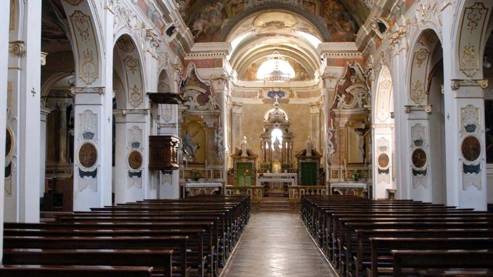 Interno di una chiesa barocca con lunga navata centrale, banchi in legno su entrambi i lati e pavimento a piastrelle diagonali. Volta affrescata e riccamente decorata con stucchi, archi laterali e altare maggiore illuminato in fondo.