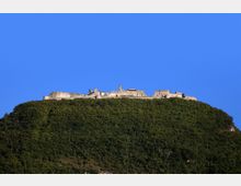 Fortezza in pietra con mura e bastioni arroccata sulla sommità di una collina completamente boscosa. Il complesso fortificato si staglia contro un cielo azzurro limpido.
