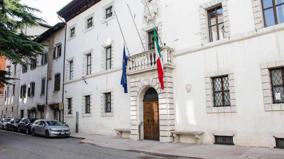 Facciata di un edificio istituzionale in intonaco bianco affacciato su una strada, con portale ad arco in pietra, balconcino e finestre incorniciate. Davanti all’ingresso sono visibili le bandiere italiana e dell’Unione Europea, con auto parcheggiate lungo il marciapiede e alberi sul lato sinistro.