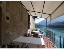 Terrazza coperta con pavimento in cotto e ringhiera in ferro battuto, arredata con tavoli e sedie vicino al muro in pietra di un edificio. Affaccia su un lago circondato da colline e montagne boscose.