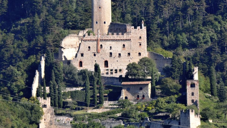 Castello medievale in pietra su un pendio boscoso, con alta torre centrale e mura merlate. In primo piano si vedono terrazzamenti e strutture fortificate minori circondate da cipressi e vegetazione.