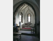 Interno di una piccola cappella in stile gotico, con volta a costoloni e arco in pietra che incornicia l’abside. Sul fondo si vedono un altare in pietra rialzato su gradini con tappeto rosso e strette finestre ad arco che lasciano filtrare luce naturale.