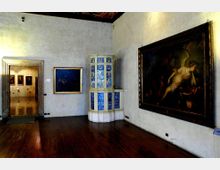 Sala museale con pavimento in legno scuro e pareti intonacate chiare, con grandi dipinti incorniciati appesi alle pareti. Al centro si trova una stufa in maiolica bianca e blu, e a sinistra un portale conduce a una stanza adiacente con altre opere esposte.