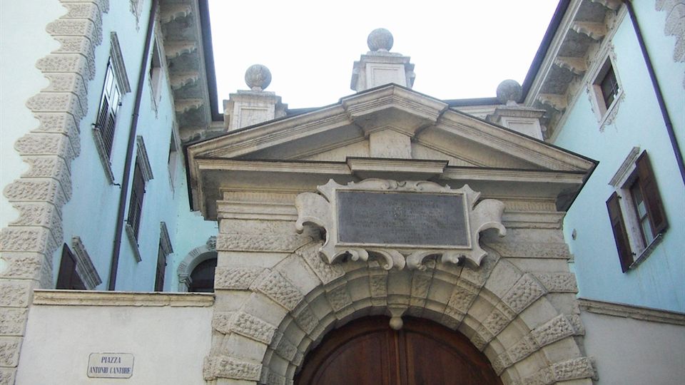 Ingresso monumentale in pietra di un palazzo storico, con grande arco bugnato, portale in legno e timpano triangolare sormontato da sfere decorative. Ai lati si vedono le facciate azzurre dell’edificio con finestre e persiane, e un cartello stradale su un muro chiaro.