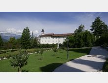 Edificio storico intonacato di bianco con tetto in coppi e un campanile centrale, immerso in un ampio giardino con prato curato, alberi e un lampione. In primo piano si vede un vialetto chiaro che costeggia l’area verde, con montagne sullo sfondo.