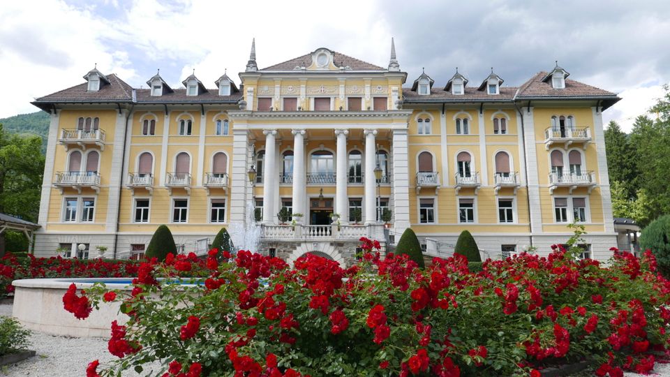 Grande edificio storico in stile neoclassico, con facciata gialla, tetto a falde con abbaini e un portico centrale con alte colonne bianche. Davanti si vede un giardino con aiuole di rose rosse, siepi potate e una fontana, con alberi e colline sullo sfondo.