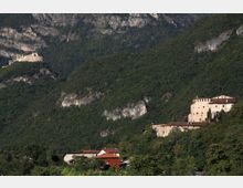 Veduta di una valle montana con pendii boscosi e pareti rocciose, con un piccolo borgo ai piedi del versante. Sul fianco della collina si distinguono edifici in pietra in stile fortificato, tra cui un complesso con torre e mura e una struttura isolata più in alto.