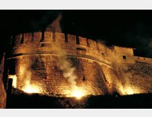 Veduta notturna di una fortezza medievale in pietra con bastione semicircolare e merlature, illuminata da luci dal basso. Fumo e fiamme alla base delle mura creano un’atmosfera di scena all’aperto.