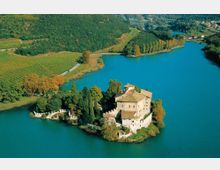 Veduta aerea di un castello con mura di cinta su una piccola penisola boscosa che si protende in un lago turchese. Sullo sfondo si vedono colline verdi, vigneti e una strada che costeggia la riva.