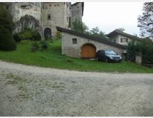 Cortile rurale con strada sterrata in primo piano e un piccolo edificio in pietra con tetto spiovente e portone ad arco. Sullo sfondo si vede una grande struttura in pietra simile a un castello e alcune case, circondate da prato e alberi.