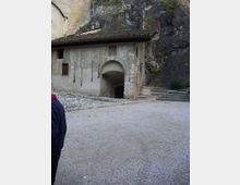 Edificio in pietra e intonaco addossato a una parete rocciosa, con tetto in coppi e un grande arco d’ingresso che conduce a un passaggio interno. Davanti si apre un cortile in ghiaia con una breve scalinata in pietra sul lato destro.