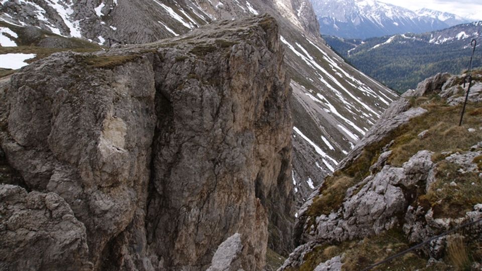 Veduta alpina con pareti rocciose ripide e cresta frastagliata con chiazze di neve, sopra un canalone che scende verso una valle boscosa. In primo piano si vede un sentiero su roccia con cavo di sicurezza lungo il bordo del dirupo.