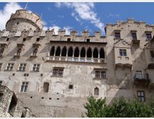 Facciata di un castello medievale in pietra chiara vista dal basso, con merlature, torre cilindrica e finestre irregolari. Al centro spicca una loggia con archi e colonnine sotto un tetto sporgente, sullo sfondo cielo azzurro con nuvole.