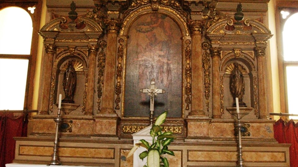 Altare di una chiesa con grande pala d’altare lignea intagliata e dorata, nicchie laterali con statue e croce in sommità. Davanti, mensa coperta da tovaglia bianca con candelieri e vasi di fiori, affiancata da finestre ad arco.