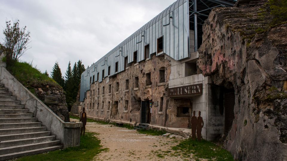 Facciata di una struttura storica scavata nella roccia, con file di piccole aperture rettangolari e un ingresso incassato, sovrastata da una copertura moderna in metallo. Un sentiero sterrato conduce all’edificio tra pareti rocciose, con una scalinata in cemento sul lato sinistro e alberi sullo sfondo.
