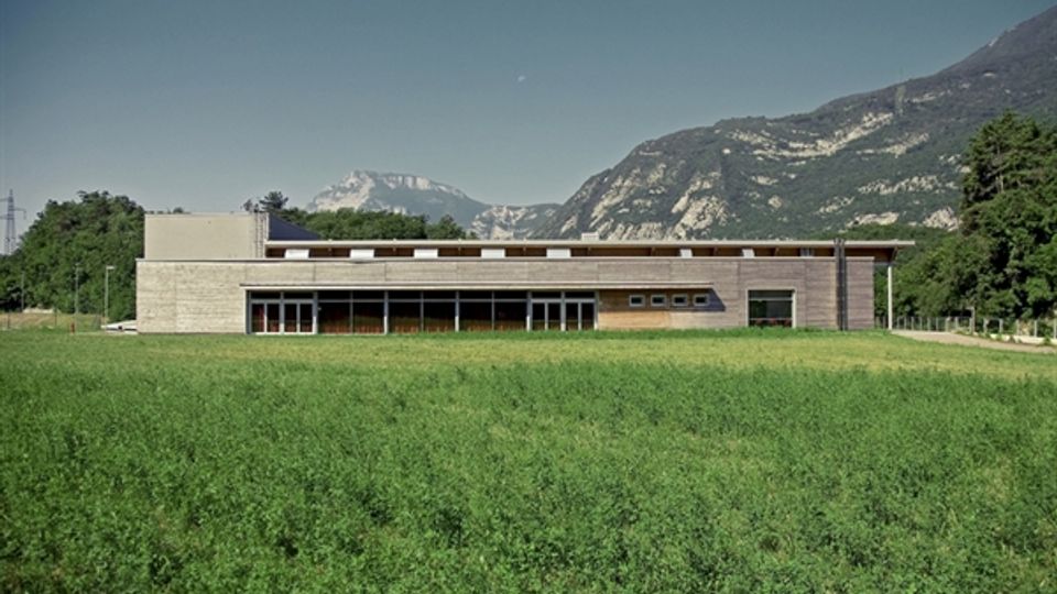 Edificio moderno a un piano con facciata in legno e pietra e ampie vetrate, situato ai margini di un grande prato verde. Sullo sfondo si vedono montagne rocciose e alberi sotto un cielo sereno.
