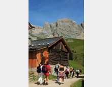 Sentiero sterrato di montagna accanto a una baita in legno e pietra con tetto in ardesia, in un prato alpino. Sullo sfondo si innalzano alte pareti rocciose grigie sotto un cielo limpido.