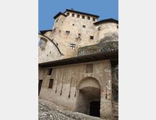 Veduta dal basso di un castello in pietra e intonaco con facciate alte e irregolari, piccole finestre e tetti sporgenti, costruito su uno sperone roccioso. In primo piano si vede un edificio d’accesso con grande arco d’ingresso e pavimentazione in ciottoli.