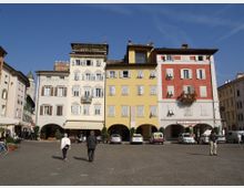 Piazza lastricata con file di edifici storici a più piani dai colori pastello (bianco, giallo e rosso), con finestre a persiane e portici ad archi al piano terra. Negozi e un’insegna “Hotel Venezia” si affacciano sulla piazza, con alcune auto parcheggiate davanti.