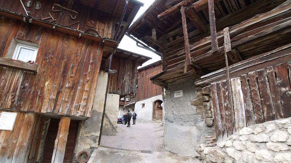 Stretto vicolo di un borgo alpino con case rustiche in legno e pietra, con balconi e travi a vista che sporgono sopra la strada. Il percorso in leggera salita conduce a un’area più aperta tra edifici in legno e intonaco.