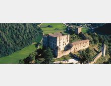 Veduta aerea di un castello medievale in pietra con torri e mura merlate, situato su un rilievo tra campi coltivati e un’area boschiva. Il complesso comprende un grande edificio principale e un lungo corpo fortificato collegato a una torre isolata.