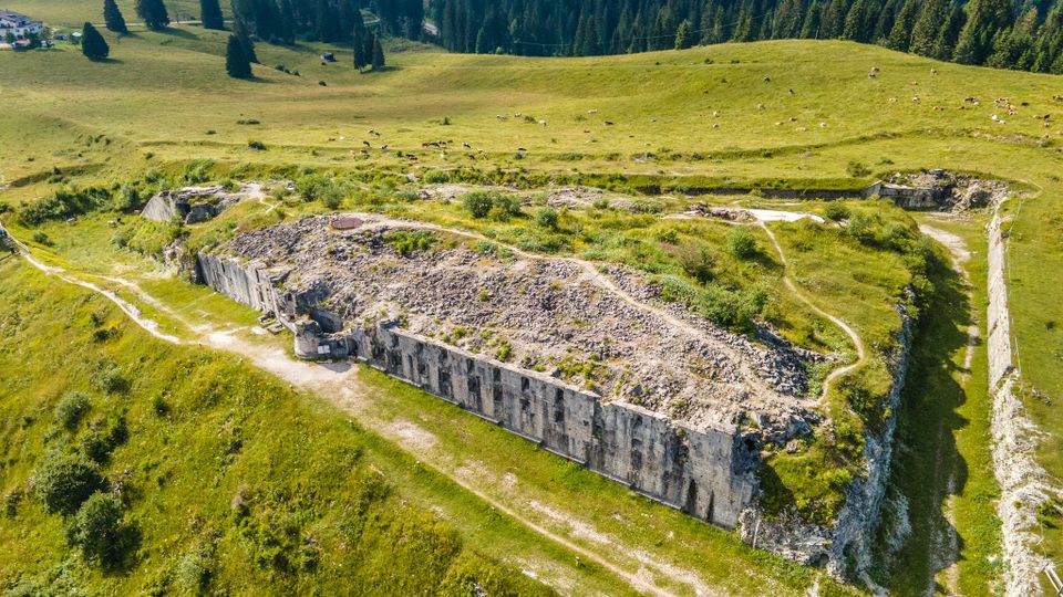 Veduta aerea di un altopiano erboso con i resti di una fortificazione in cemento, caratterizzata da lunghi muri parzialmente crollati e sentieri sterrati intorno. Sullo sfondo si estendono prati ondulati e una fitta foresta di conifere.