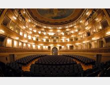 Interno di un teatro all’italiana con platea di poltrone scure e diversi ordini di palchi curvi illuminati da appliques. Soffitto a cupola affrescato e decorazioni dorate lungo le balconate e le pareti.