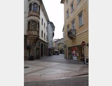 Strada pedonale acciottolata in un centro storico, fiancheggiata da edifici a più piani con facciate chiare, finestre ad arco e un balcone in ferro battuto. In fondo si vede una piccola piazza con ombrelloni di un locale e altri palazzi lungo la via.
