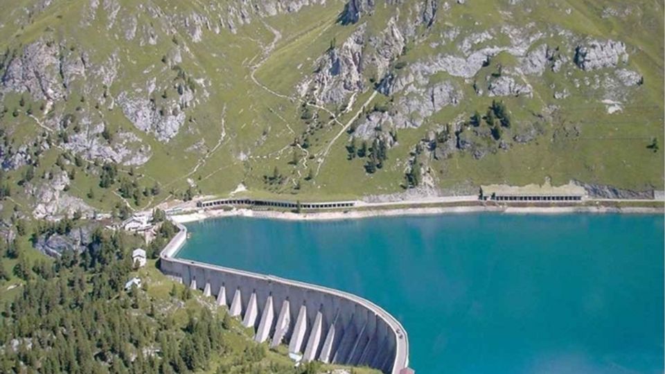Diga curva in cemento che trattiene un lago artificiale dall’acqua turchese, ai piedi di pendii montani erbosi con affioramenti rocciosi. In primo piano si vede una strada sul coronamento della diga e, più in basso, un piccolo complesso di edifici e bosco.