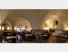 Ampia sala interna con soffitto a volte a crociera in stile gotico e grandi archi sostenuti da pilastri in pietra, illuminata da applique a parete. Nello spazio sono disposti tavoli e sedie, con finestre ad arco sul fondo e un affresco decorativo su una parete.