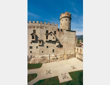 Veduta esterna di un castello in pietra con mura merlate e una torre cilindrica, con piccole finestre e volumi sporgenti sulla facciata. In primo piano si vede un cortile con vialetti di ghiaia, aiuole verdi e alcune strutture basse quadrate.
