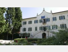 Edificio storico a tre piani con facciata bianca, finestre con scuri verdi e frontone centrale curvilineo con balcone e bandiere. Davanti si trovano un giardino con siepi e fiori, alberi alti e una scala esterna che conduce all’ingresso ad arco.