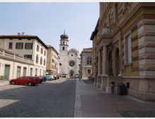 Strada urbana lastricata fiancheggiata da edifici storici, con auto parcheggiate lungo il lato sinistro e un grande palazzo con portici sul lato destro. In fondo si vede una chiesa in pietra con rosone centrale e campanile con cupola a bulbo.