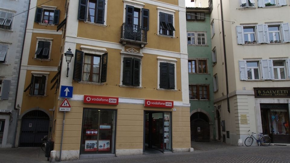 Edificio storico a più piani con facciata ocra e persiane scure, affacciato su una piazza lastricata; al piano terra ci sono vetrine di negozi e un lampione stradale. Ai lati si vedono altri palazzi con persiane chiare e un passaggio ad arco tra gli edifici.