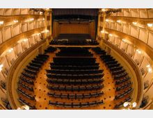 Interno di un teatro con platea e palchi laterali multipli decorati in stile classico, illuminati da applique dorate. Inquadratura dall’alto verso il palcoscenico con sipario e file di poltrone disposte a semicerchio su pavimento in legno.