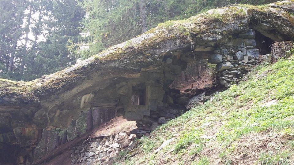 Sporgenza rocciosa coperta di muschio su un pendio boschivo, con una piccola cavità e muri a secco in pietra che formano un riparo sotto la roccia. Attorno si vedono alberi di conifere e terreno in salita con erba e pietre sparse.