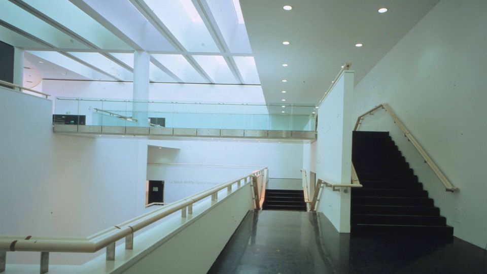 Interno di un edificio moderno con atrio a doppia altezza, pareti bianche e pavimento scuro lucido. Una rampa con corrimano conduce a un ballatoio con parapetto in vetro, illuminato da un lucernario a travi e faretti a soffitto, con scala laterale in fondo.