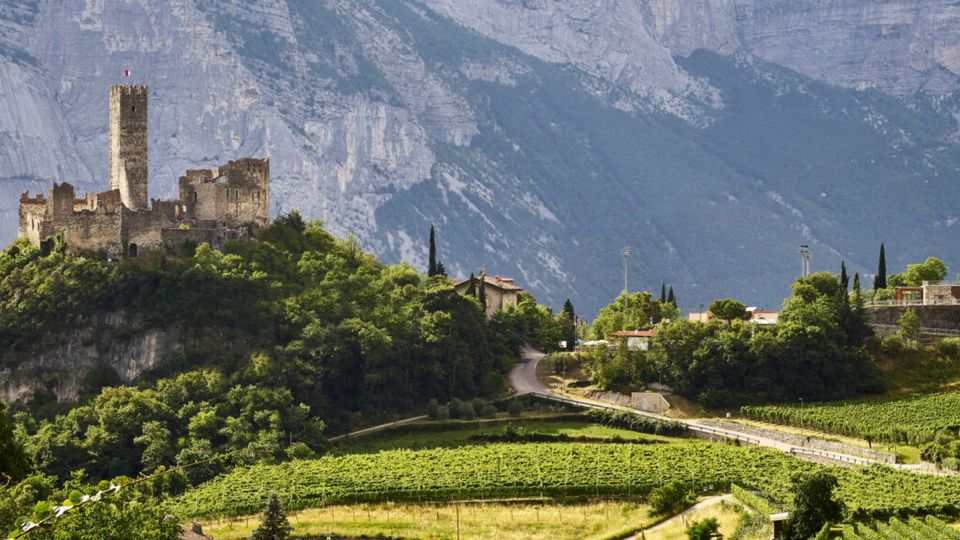 Rovine di un castello medievale con alta torre in pietra su una collina boscosa, con vigneti terrazzati e una strada che serpeggia nella valle sottostante. Sullo sfondo si vedono ripide pareti montuose rocciose e alcune case sparse tra gli alberi.