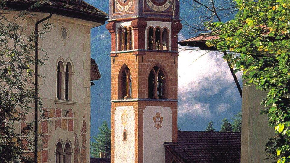 Torre dell’orologio in muratura con quadranti su più lati e coronamento merlato, affiancata da edifici storici. Sullo sfondo si vedono montagne alpine e alberi ai margini della strada del centro abitato.
