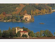 Veduta aerea di un lago con due piccole penisole alberate, ciascuna con una villa storica in muratura e tetti in coppi. Sullo sfondo si vedono colline con vigneti e filari di pioppi lungo la riva.