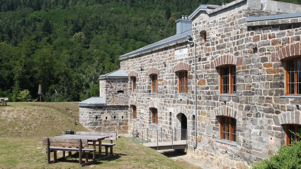Edificio storico in pietra con finestre ad arco e tetto in metallo, affacciato su un prato con tavolo e panche in legno. Sullo sfondo si vede un fitto bosco che copre il pendio della montagna.