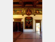 Interno di una sala storica con soffitto a cassettoni in legno intagliato e fregio decorato con figure e stemma centrale. Sulla parete bianca è appeso un grande dipinto incorniciato, affiancato da due portali con cornici in pietra e pavimento in terrazzo lucido.