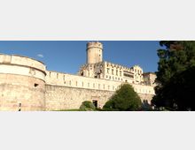 Castello in pietra con alte mura merlate e torrione cilindrico centrale, affiancato da edifici storici con finestre ad arco. In primo piano si vedono il bastione curvo, un prato con cespugli e alberi, sotto un cielo sereno.