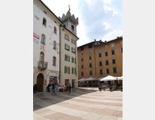 Piazza lastricata in un centro storico, circondata da edifici alti dai colori chiari con finestre e persiane, e una torre in pietra con merli sullo sfondo. Sul lato destro ci sono tavolini all’aperto sotto ombrelloni e alcune fioriere.