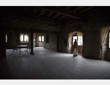 Ampia stanza interna spoglia in un edificio antico, con pavimento in assi di legno, travi a vista e muri grezzi. Diverse finestre e una porta con tende leggere lasciano entrare luce naturale; pochi arredi essenziali sono presenti in un angolo.