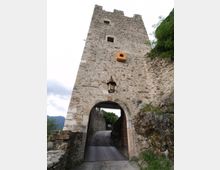 Torre in pietra di una fortificazione medievale con grande arco d’ingresso che sovrasta una strada stretta in salita. Sulla facciata si notano piccole finestre e una lanterna appesa, con mura e roccia ai lati e vegetazione circostante.