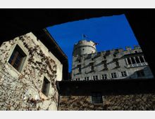 Veduta dal basso di un castello in pietra con torre cilindrica e merli, affacciato su un cortile interno. A sinistra si vede una facciata con finestre rettangolari ricoperta di rampicanti, sotto un cielo blu intenso.